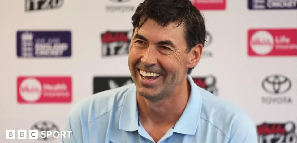 Coach smiling during press conference with logos behind